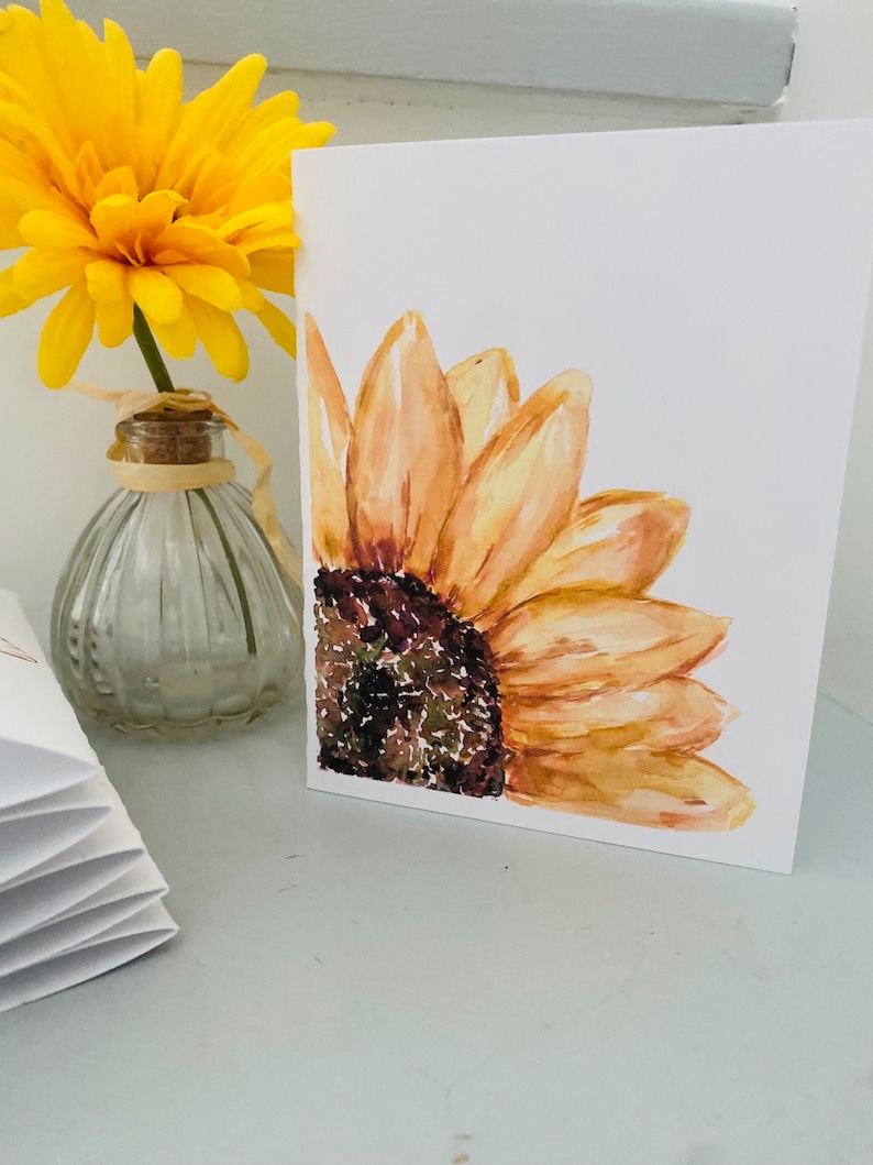 Puede incluir: Una tarjeta de felicitaci&oacute;n blanca con una pintura de acuarela de un girasol. El girasol tiene p&eacute;talos amarillos y un centro marr&oacute;n oscuro. Una flor amarilla en un jarr&oacute;n de cristal est&aacute; en el fondo.