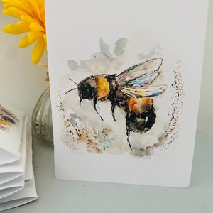 Puede incluir: Tarjeta de felicitaci&oacute;n de acuarela con una ilustraci&oacute;n detallada de un abejorro en vuelo. La abeja est&aacute; representada con rayas negras y amarillas, y la obra de arte tiene un estilo suave y pict&oacute;rico. Una flor amarilla en un jarr&oacute;n est&aacute; en el fondo.