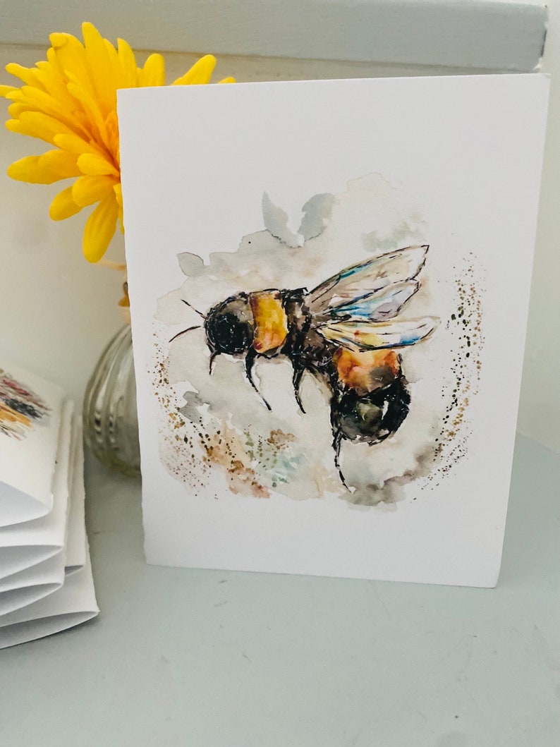 Puede incluir: Tarjeta de felicitaci&oacute;n de acuarela con una ilustraci&oacute;n detallada de un abejorro en vuelo. La abeja est&aacute; representada con rayas negras y amarillas, y la obra de arte tiene un estilo suave y pict&oacute;rico. Una flor amarilla en un jarr&oacute;n est&aacute; en el fondo.