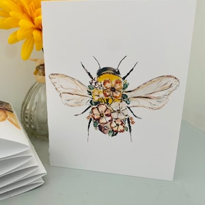 Puede incluir: Una tarjeta blanca con una ilustraci&oacute;n en acuarela de una abeja adornada con flores. La abeja tiene un cuerpo amarillo y negro, con delicadas alas y acentos florales en rosa, marr&oacute;n y amarillo.