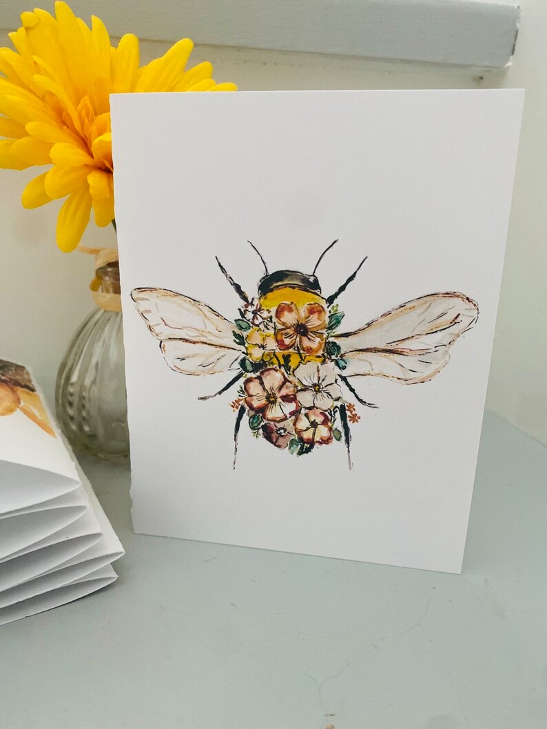 Puede incluir: Una tarjeta blanca con una ilustraci&oacute;n en acuarela de una abeja adornada con flores. La abeja tiene un cuerpo amarillo y negro, con delicadas alas y acentos florales en rosa, marr&oacute;n y amarillo.