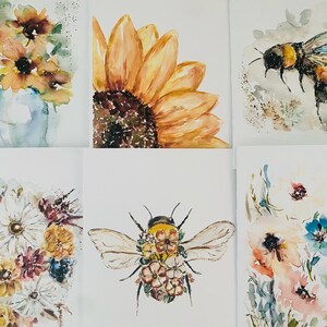 Puede incluir: Pinturas a la acuarela de arreglos florales e insectos. La obra de arte incluye un girasol, una abeja y ramos de flores en tonos amarillos, naranjas, azules y rosas. Las pinturas est&aacute;n sobre un fondo blanco.