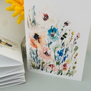 Puede incluir: Tarjeta de felicitaci&oacute;n de acuarela con un dise&ntilde;o floral con flores rosas, azules y blancas. La tarjeta se muestra junto a una flor amarilla y una pila de tarjetas en blanco. La obra de arte tiene un estilo suave y pict&oacute;rico.