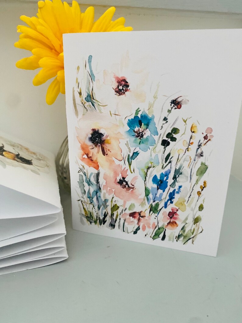 Puede incluir: Tarjeta de felicitaci&oacute;n de acuarela con un dise&ntilde;o floral con flores rosas, azules y blancas. La tarjeta se muestra junto a una flor amarilla y una pila de tarjetas en blanco. La obra de arte tiene un estilo suave y pict&oacute;rico.