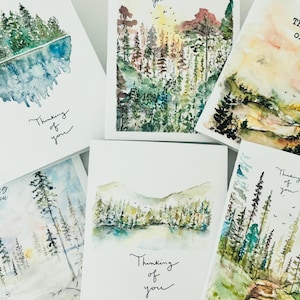 Può includere: Una collezione di cinque biglietti d'auguri bianchi con illustrazioni paesaggistiche ad acquerello di foreste, montagne e laghi. Ogni biglietto mostra il testo scritto a mano Thinking of you in inchiostro nero, raffigurante varie scene naturali con toni verdi, blu e gialli.