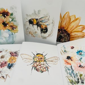 Puede incluir: Colecci&oacute;n de tarjetas de felicitaci&oacute;n de acuarela con dise&ntilde;os florales e insectos. Las tarjetas muestran girasoles, abejas y flores silvestres en tonos amarillos, azules y rosas. La obra de arte tiene un estilo suave y art&iacute;stico.