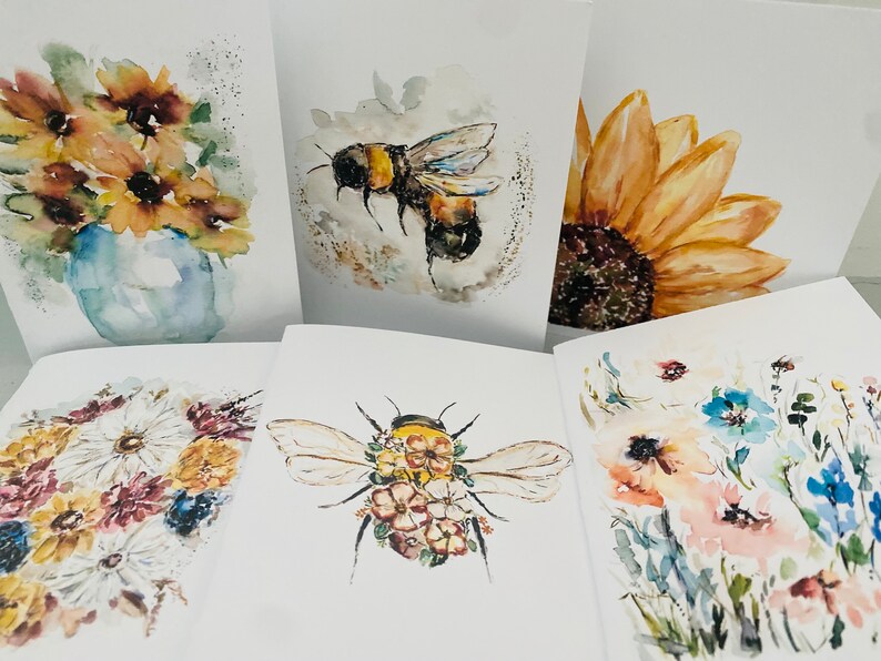 Puede incluir: Colecci&oacute;n de tarjetas de felicitaci&oacute;n de acuarela con dise&ntilde;os florales e insectos. Las tarjetas muestran girasoles, abejas y flores silvestres en tonos amarillos, azules y rosas. La obra de arte tiene un estilo suave y art&iacute;stico.