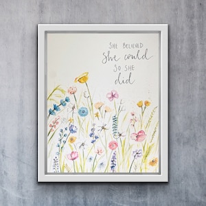 Op de afbeelding: Een aquarel van een veld met wilde bloemen met de quote "She believed she could so she did" geschreven in zwarte cursieve letters.
