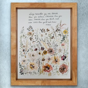 May include: A framed watercolor print featuring a floral design in soft hues of peach, cream, and brown, with butterflies. The artwork includes the quote: "always remember you are BRAVER than you believe..." in black script. The frame is made of light-colored wood.