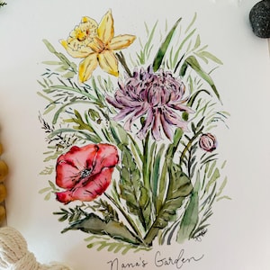 May include: Watercolor painting of a bouquet of flowers, including a yellow daffodil, a pink chrysanthemum, and a red poppy. The flowers are surrounded by green leaves and stems. The painting is signed "Nana's Garden" in the bottom right corner.