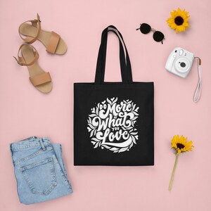 Peut inclure: Sac fourre-tout noir avec inscription blanche "Do More of What You Love" entourée de motifs floraux. Le sac est sur un fond rose avec une paire de sandales, un appareil photo, des lunettes de soleil et des tournesols.
