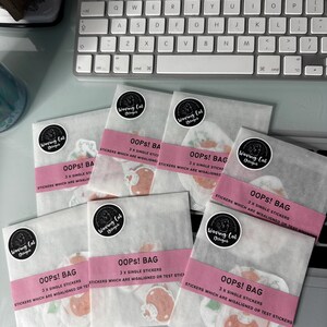 Può includere: Sei sacchetti di carta bianca con etichette rosa che recitano "Oops! Bag" e "3 x adesivi singoli che sono disallineati o adesivi di prova". Ogni sacchetto ha un logo in bianco e nero che dice "Weaving Cat Designs".