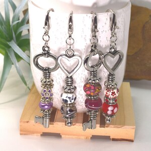 May include: Four silver-toned metal key-shaped charms featuring lobster claw clasps. Each charm is decorated with various beads, including purple, white, black, pink, and red patterns, along with small silver spacers and heart or oval tops.
