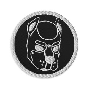 以下が含まれることがあります： 犬の口輪のデザインが施された黒と白の刺繍パッチ。口輪は白で縁取られ、黒の背景になっています。
