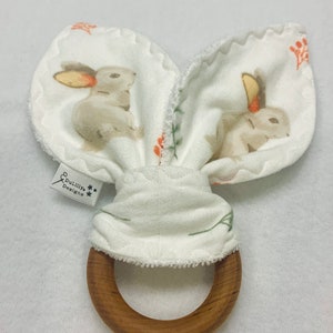 May include: A white fabric bunny rabbit teether with a wooden ring. The fabric has a small floral pattern and a small tag that says "Dudlily Designs".