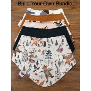 May include: A stack of five triangular baby bibs with the text Build Your Own Bundle. The bibs feature various patterns including a cream background with woodland animals, a solid dark charcoal, a light pink with squirrels, a solid tan, and a white with acorns.
