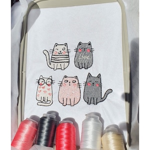 Könnte beinhalten: Fünf gestickte Katzenmotive auf einem weißen Stoff mit dem Wort "Love" auf einer der Katzen gestickt. Die Katzen sind in verschiedenen Farben, darunter Schwarz, Weiß, Rosa und Grau. Es gibt sechs Spulen Stickgarn in verschiedenen Farben, darunter Rot, Schwarz, Weiß und Beige.