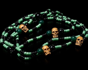 May include: A long beaded necklace featuring alternating segments of small mint green and glossy black seed beads. The strand is accented with smooth, dark green round beads and four carved, beige-colored skull beads spaced evenly throughout.