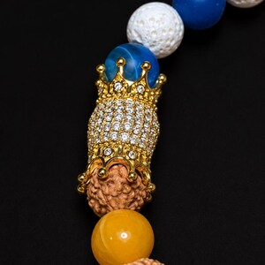 Pu&ograve; includere: Un braccialetto di perline con perle di lava bianca porosa, perle sferiche lisce blu e gialle e semi di rudraksha marroni testurizzati. Una perla centrale in metallo dorato a forma di corona &egrave; incastonata con piccoli cristalli trasparenti.
