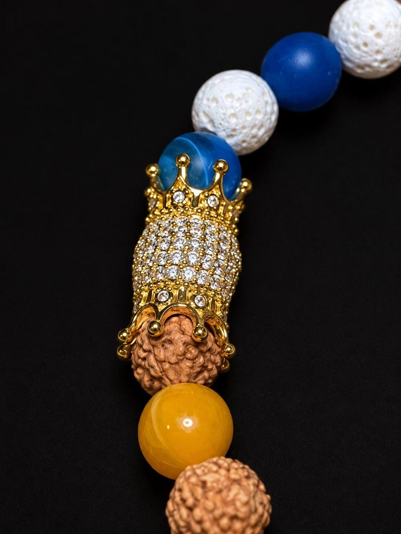 Pu&ograve; includere: Un braccialetto di perline con perle di lava bianca porosa, perle sferiche lisce blu e gialle e semi di rudraksha marroni testurizzati. Una perla centrale in metallo dorato a forma di corona &egrave; incastonata con piccoli cristalli trasparenti.