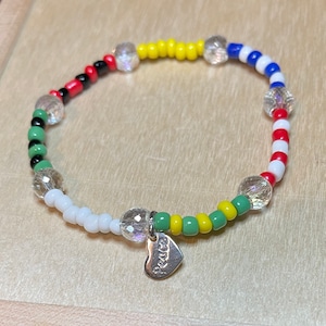 May include: A colorful beaded bracelet with a heart charm that says "peace". The bracelet features alternating beads of red, white, blue, yellow, and green.
