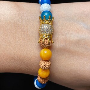 Pu&ograve; includere: Un braccialetto di perline con un ciondolo centrale a forma di corona color oro incastonato con cristalli trasparenti, affiancato da perline rudraksha marroni testurizzate, perline gialle lisce, perline blu e perline bianche craquel&eacute;.
