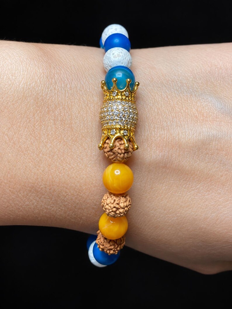 Pu&ograve; includere: Un braccialetto di perline con un ciondolo centrale a forma di corona color oro incastonato con cristalli trasparenti, affiancato da perline rudraksha marroni testurizzate, perline gialle lisce, perline blu e perline bianche craquel&eacute;.