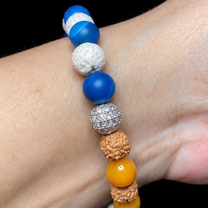 Pu&ograve; includere: Un braccialetto di perline composto da una sequenza di sfere blu lisce, pietre laviche bianche porose, una singola perla color argento incastonata con cristalli trasparenti e semi di rudraksha marroni testurizzati, intervallati da perle gialle.