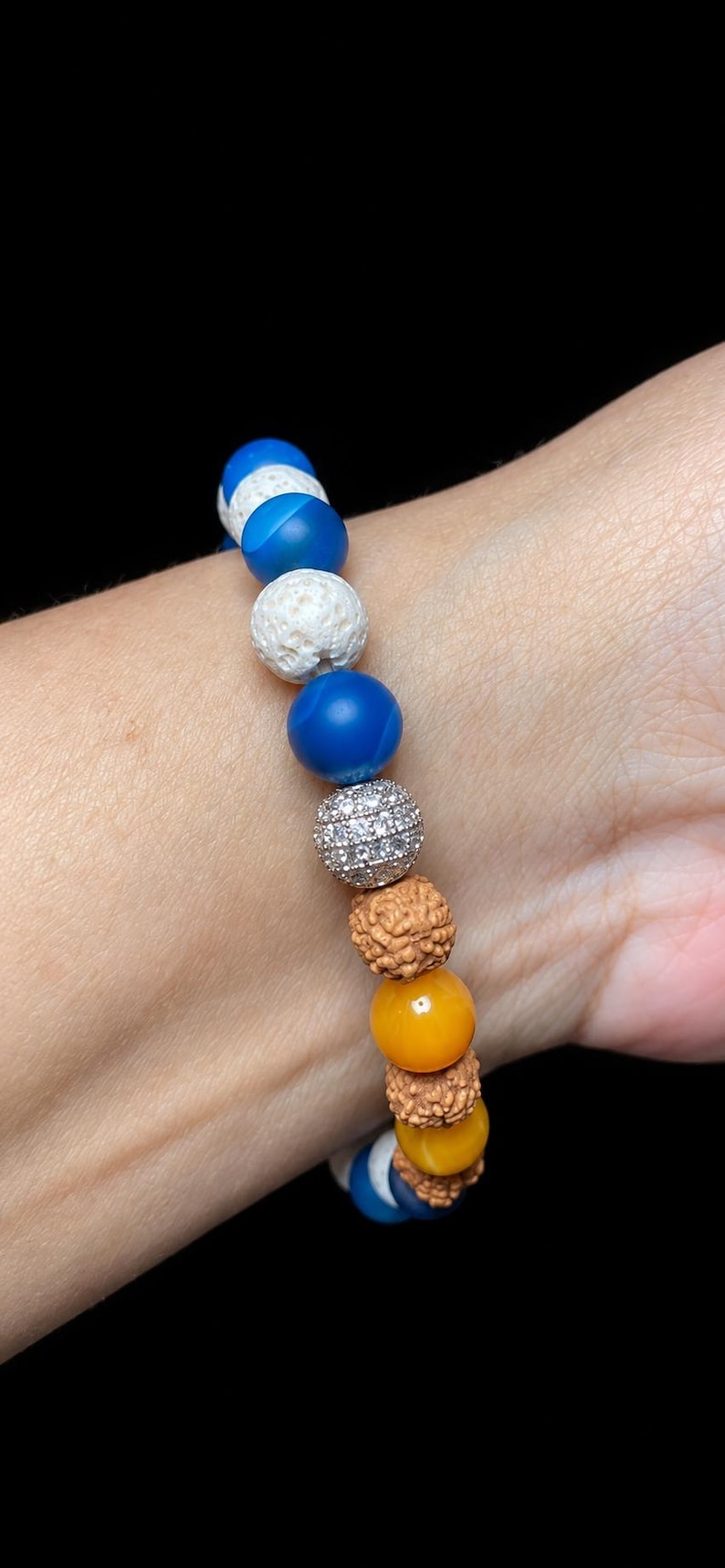 Pu&ograve; includere: Un braccialetto di perline composto da una sequenza di sfere blu lisce, pietre laviche bianche porose, una singola perla color argento incastonata con cristalli trasparenti e semi di rudraksha marroni testurizzati, intervallati da perle gialle.