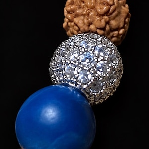 Pu&ograve; includere: Un braccialetto di perline con una sequenza di perline rudraksha marroni testurizzate, una perlina gialla liscia, una perlina sferica color argento incastonata con cristalli trasparenti, una perlina blu reale liscia e una perlina bianca porosa su sfondo nero.