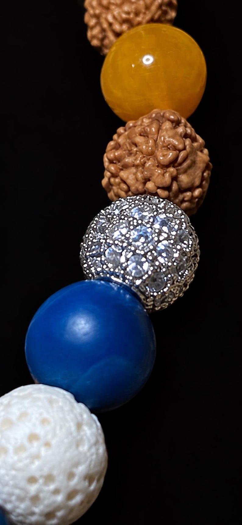Pu&ograve; includere: Un braccialetto di perline con una sequenza di perline rudraksha marroni testurizzate, una perlina gialla liscia, una perlina sferica color argento incastonata con cristalli trasparenti, una perlina blu reale liscia e una perlina bianca porosa su sfondo nero.