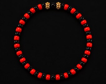 Può includere: Un braccialetto circolare di perline con perline cilindriche rosse e nere alternate disposte in un motivo ripetitivo. Due perline decorative metalliche color oro con dettagli strutturati complessi sono posizionate al centro superiore.