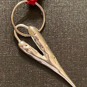 May include: A silver heart-shaped keychain with a textured surface and a pointed tip. The keychain is attached to a silver ring and a red leather cord.