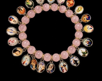 Puede incluir: Una pulsera circular con cuentas de cuarzo rosa pálido y liso. Enganchados a los eslabones de metal dorado entre las cuentas hay numerosos dijes pequeños de forma ovalada, cada uno con un retrato religioso colorido.