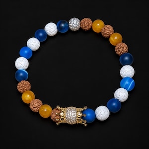 Pu&ograve; includere: Un braccialetto di perline con un mix di perline blu, bianche, gialle e marroni testurizzate. Il design include sfere blu lisce, perline di lava bianche porose, perline gialle traslucide e semi di rudraksha marroni con una perlina a corona.