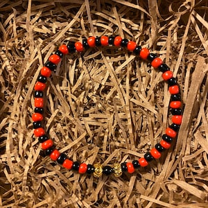 Puede incluir: Un brazalete de cuentas negras y naranjas con detalles dorados. El brazalete está hecho de pequeñas cuentas redondas y está enhebrado en un cordón delgado.