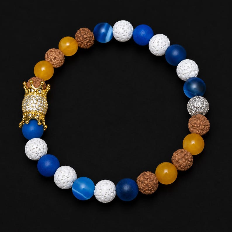 Pu&ograve; includere: Un braccialetto di perline con un mix di perline testurizzate blu, gialle, bianche e marroni. Il design include una prominente perlina cilindrica color oro incastonata con piccole pietre chiare, a forma di corona su entrambe le estremit&agrave;.