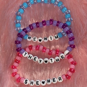 May include: Three beaded bracelets with the words "He Him", "They Them", and "She Her" spelled out with black letters and a small heart between each word.
