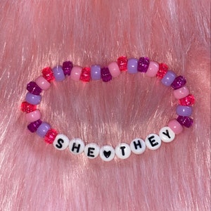 May include: A beaded bracelet with pink, purple, and red beads. The bracelet spells out "SHE &hearts; THEY" in white letters.