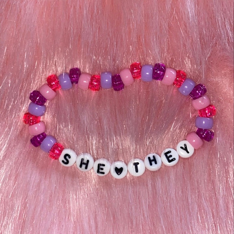 May include: A beaded bracelet with pink, purple, and red beads. The bracelet spells out "SHE &hearts; THEY" in white letters.
