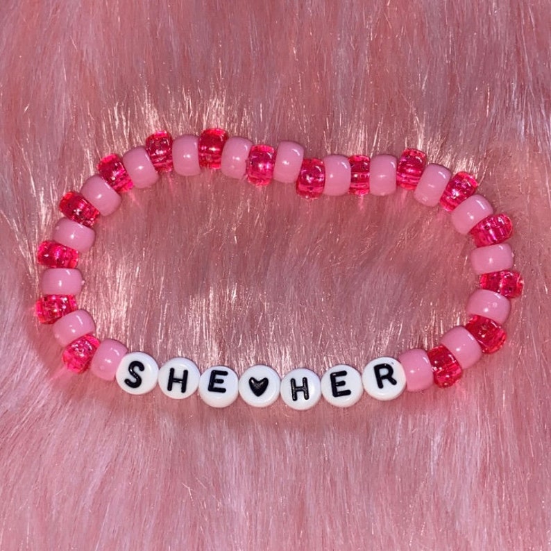 May include: Pink beaded bracelet with the words "SHE &hearts; HER" in black letters.