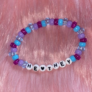 May include: A beaded bracelet with purple, blue, and clear beads. The bracelet spells out "HE &hearts; THEY" in white letters.