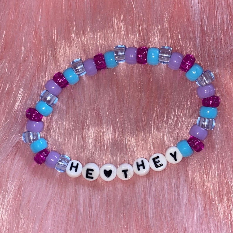 May include: A beaded bracelet with purple, blue, and clear beads. The bracelet spells out "HE &hearts; THEY" in white letters.