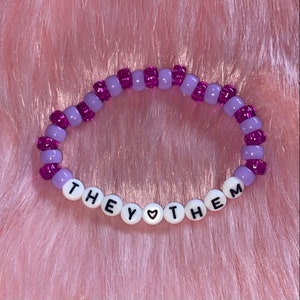 May include: Purple and lavender beaded bracelet with the words "THEY &hearts; THEM" spelled out in white beads.