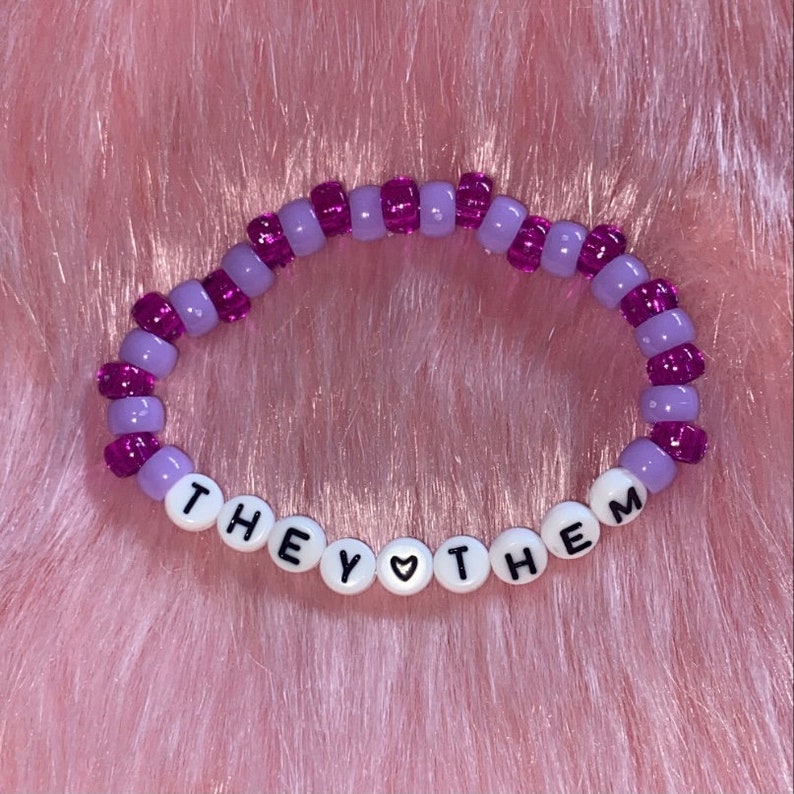 May include: Purple and lavender beaded bracelet with the words "THEY &hearts; THEM" spelled out in white beads.