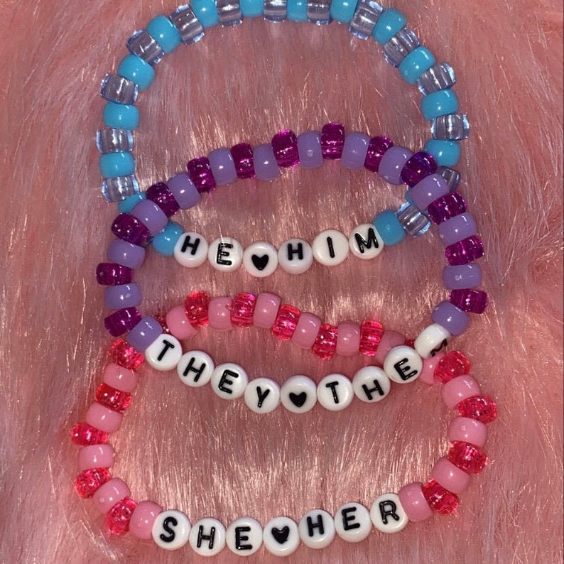 May include: Three beaded bracelets with the words "He Him", "They Them", and "She Her" spelled out with black letters and a small heart between each word.