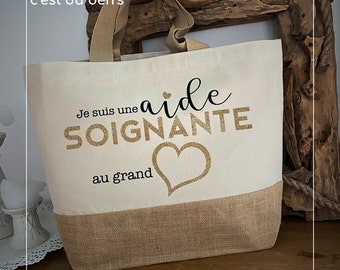 May include: Cream-colored tote bag with a burlap bottom and handles. The bag features the text "Je suis une aide SOIGNANTE au grand" in gold glitter lettering, along with a heart graphic. The text "créé par c'est du ben's" is at the top.