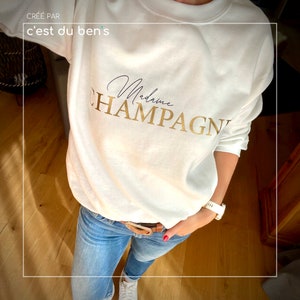 Peut inclure: Sweat-shirt blanc avec le texte "Madame Champagne" en lettres dorées. Le sweat-shirt est porté par une personne avec un jean bleu et une montre blanche.