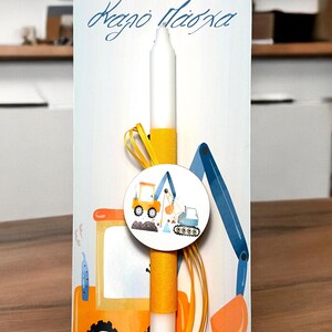 May include: A white candle with a yellow ribbon and a round sticker featuring a cartoon excavator and bulldozer. The candle is attached to a wooden stand with the text "Χαρού Πάσχα" in blue lettering.