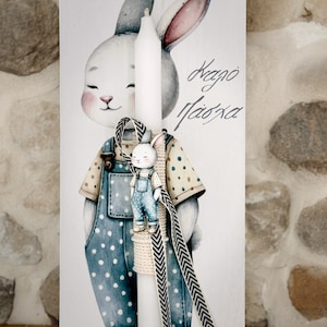 May include: White Easter candle with a bunny figurine attached. The bunny is wearing blue overalls with white polka dots and a brown and white striped ribbon. The candle is on a white background with a bunny illustration and the text "Καλο Πάσχα".